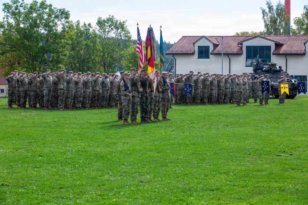 A military ceremony is taking place on a grassy parade ground, featuring soldiers in camouflage uniforms standing in formation while saluting. A color guard stands at center holding multiple flags including the United States flag and the German flag, suggesting a joint US-German military event. The setting appears to be a military installation in Germany, with a white building with red tile roof and an armored vehicle visible in the background.