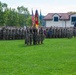 A military ceremony is taking place on a grassy parade ground, featuring soldiers in camouflage uniforms standing in formation while saluting. A color guard stands at center holding multiple flags including the United States flag and the German flag, suggesting a joint US-German military event. The setting appears to be a military installation in Germany, with a white building with red tile roof and an armored vehicle visible in the background.