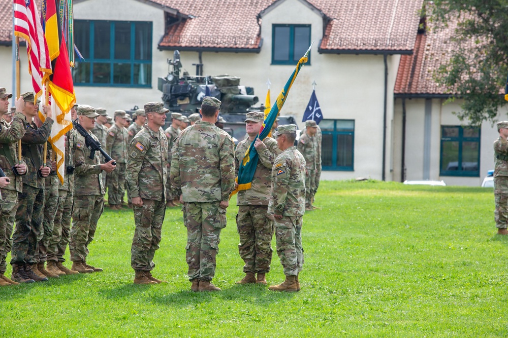 A formal U.S. Army military ceremony, likely a change of command or activation/deactivation ceremony, is taking place on a grass parade field. Several soldiers in OCP (Operational Camouflage Pattern) uniforms are centered in the frame, with one soldier passing or receiving a unit guidon (teal/green flag). Rows of soldiers stand in formation in the background, with multiple flags including U.S. and NATO flags visible.