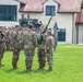 A formal U.S. Army military ceremony, likely a change of command or activation/deactivation ceremony, is taking place on a grass parade field. Several soldiers in OCP (Operational Camouflage Pattern) uniforms are centered in the frame, with one soldier passing or receiving a unit guidon (teal/green flag). Rows of soldiers stand in formation in the background, with multiple flags including U.S. and NATO flags visible.