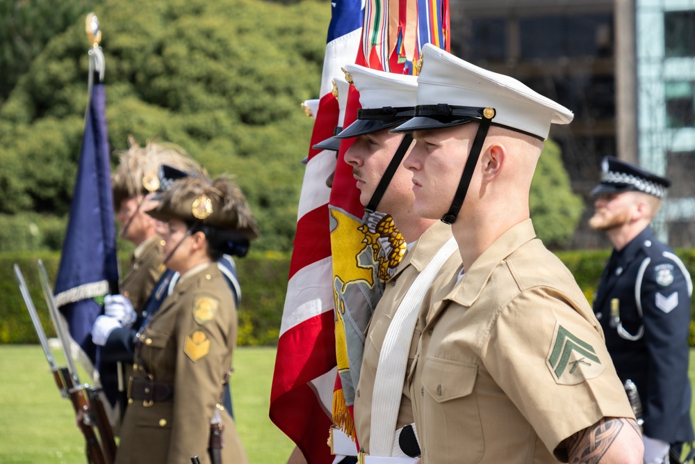 A multinational military color guard ceremony is taking place outdoors on a sunny day. U.S. Marines in khaki service uniforms and white covers are prominently featured carrying the American flag alongside personnel from what appear to be British/Commonwealth military forces in olive dress uniforms. A police officer in a checkered-band cap is also visible in the background.