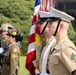 A multinational military color guard ceremony is taking place outdoors on a sunny day. U.S. Marines in khaki service uniforms and white covers are prominently featured carrying the American flag alongside personnel from what appear to be British/Commonwealth military forces in olive dress uniforms. A police officer in a checkered-band cap is also visible in the background.