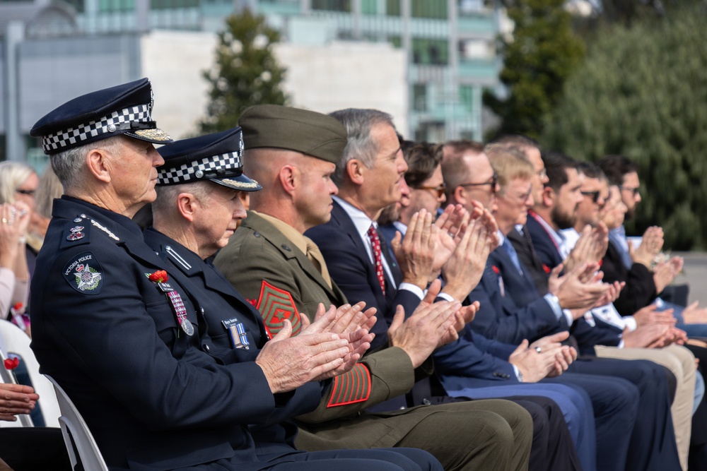 A formal commemorative ceremony featuring uniformed officials and civilians seated in a row, all applauding. The attendees include Victoria Police officers in dark uniforms with checkered caps, a military personnel in olive/khaki uniform with red sergeant chevrons, and multiple civilians in business attire. Several attendees are wearing red poppy pins, suggesting this is an ANZAC Day or Remembrance Day event in Australia.
