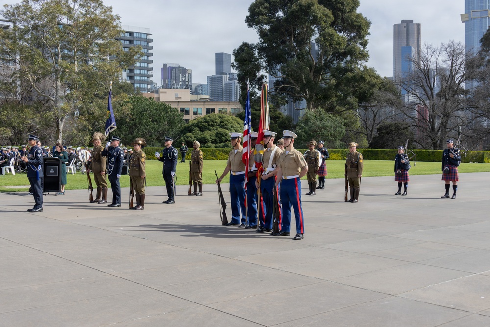 A formal military ceremony is taking place on a paved plaza, likely at a war memorial or commemorative site in an Australian city, identifiable by the Melbourne skyline in the background. Multiple military personnel from different services and nations are present, including US Marines carrying the American flag, Australian Army soldiers in khaki uniforms, and Scottish-style bagpipers in tartan kilts. The event appears to be a joint commemorative parade or memorial service.
