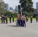 A formal military ceremony is taking place on a paved plaza, likely at a war memorial or commemorative site in an Australian city, identifiable by the Melbourne skyline in the background. Multiple military personnel from different services and nations are present, including US Marines carrying the American flag, Australian Army soldiers in khaki uniforms, and Scottish-style bagpipers in tartan kilts. The event appears to be a joint commemorative parade or memorial service.