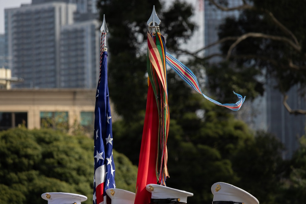 The image shows a military color guard ceremony with personnel in white dress covers (caps) holding flag staffs. A United States flag is visible on the left, while the center staff displays what appears to be a unit guidon or streamer-adorned battle color with multiple campaign streamers flowing from the finial. The setting appears to be an outdoor urban environment with trees and modern buildings visible in the background.