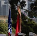 The image shows a military color guard ceremony with personnel in white dress covers (caps) holding flag staffs. A United States flag is visible on the left, while the center staff displays what appears to be a unit guidon or streamer-adorned battle color with multiple campaign streamers flowing from the finial. The setting appears to be an outdoor urban environment with trees and modern buildings visible in the background.