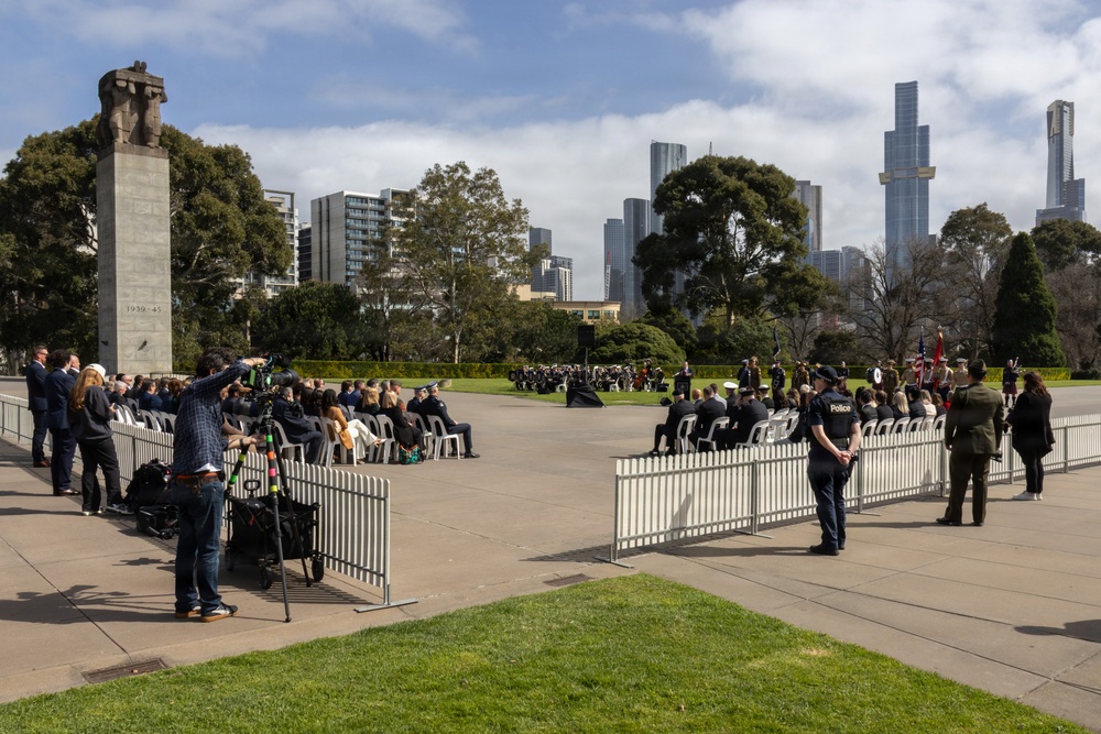 A formal commemorative ceremony is taking place at what appears to be the Shrine of Remembrance in Melbourne, Australia, identifiable by the stone monument inscribed with '1939-45' and the recognizable city skyline in the background. Attendees are seated in rows of chairs on a large paved forecourt, with police officers, military personnel, and media crews present. Temporary crowd-control barriers separate different sections of the ceremony area.