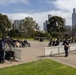A formal commemorative ceremony is taking place at what appears to be the Shrine of Remembrance in Melbourne, Australia, identifiable by the stone monument inscribed with '1939-45' and the recognizable city skyline in the background. Attendees are seated in rows of chairs on a large paved forecourt, with police officers, military personnel, and media crews present. Temporary crowd-control barriers separate different sections of the ceremony area.