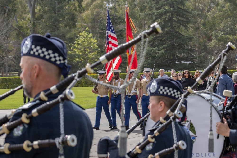 A formal military and ceremonial procession is taking place outdoors, featuring a U.S. Marine Corps color guard carrying the American flag and Marine Corps flag. In the foreground, bagpipers wearing dark blue uniforms with black-and-white checkered caps — consistent with a police pipe band — are playing their instruments. A crowd of civilian attendees follows behind the color guard, suggesting a memorial service, funeral, or official ceremony.