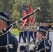 A formal military and ceremonial procession is taking place outdoors, featuring a U.S. Marine Corps color guard carrying the American flag and Marine Corps flag. In the foreground, bagpipers wearing dark blue uniforms with black-and-white checkered caps — consistent with a police pipe band — are playing their instruments. A crowd of civilian attendees follows behind the color guard, suggesting a memorial service, funeral, or official ceremony.