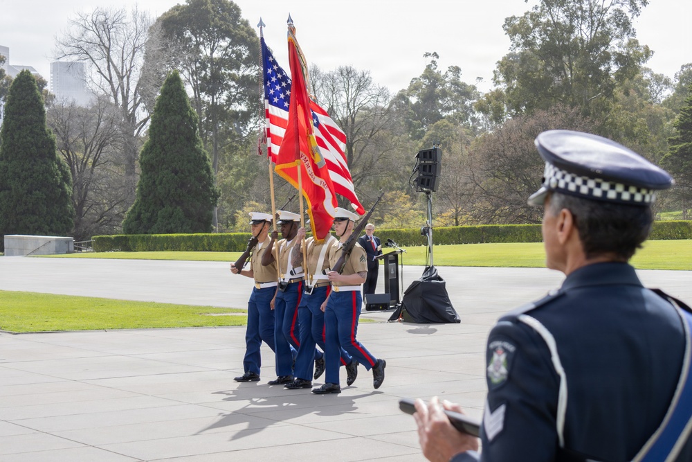 A U.S. Marine Corps color guard is marching in a formal outdoor ceremony, carrying the American flag and the Marine Corps flag. An Australian police officer in a distinctive checkered-band cap observes the procession from the foreground. The setting appears to be a memorial or civic plaza, likely in Australia, suggesting a joint or commemorative military event.