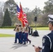 A U.S. Marine Corps color guard is marching in a formal outdoor ceremony, carrying the American flag and the Marine Corps flag. An Australian police officer in a distinctive checkered-band cap observes the procession from the foreground. The setting appears to be a memorial or civic plaza, likely in Australia, suggesting a joint or commemorative military event.