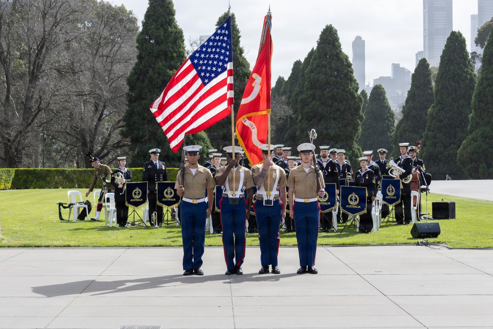 A military color guard ceremony is taking place outdoors, featuring U.S. Marines presenting the American flag and a red Marine Corps flag. Behind them, a military band with banners reading 'AUSTRALIA' performs, suggesting a joint U.S.-Australian military event. The setting appears to be a formal memorial or ceremonial ground with a city skyline visible in the background.