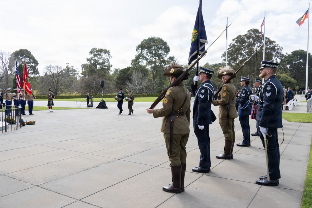 A formal military and law enforcement honor guard ceremony is taking place at an outdoor memorial or ceremonial ground, likely in Australia. Personnel from multiple services including the Australian Army, Australian Federal Police, and what appears to be a state police force are standing at attention holding rifles and a dark flag. In the background, US Marine Corps color guards and a bagpiper are also visible, suggesting a joint Australia-US commemorative event.