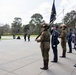 A formal military and law enforcement honor guard ceremony is taking place at an outdoor memorial or ceremonial ground, likely in Australia. Personnel from multiple services including the Australian Army, Australian Federal Police, and what appears to be a state police force are standing at attention holding rifles and a dark flag. In the background, US Marine Corps color guards and a bagpiper are also visible, suggesting a joint Australia-US commemorative event.