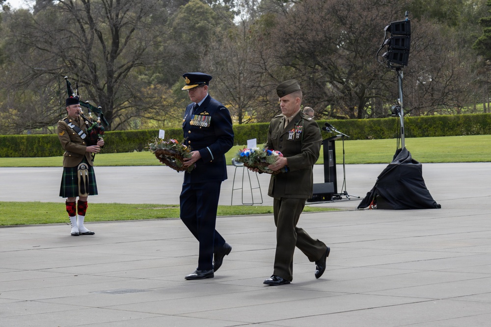 A formal military wreath-laying ceremony is taking place outdoors on a paved plaza, likely at a war memorial or commemorative site. Two senior military officers — one in a dark blue air force uniform and one in an olive/green marine uniform — are carrying floral wreaths. A bagpiper in traditional Scottish military dress with tartan kilt accompanies the procession.