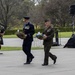 A formal military wreath-laying ceremony is taking place outdoors on a paved plaza, likely at a war memorial or commemorative site. Two senior military officers — one in a dark blue air force uniform and one in an olive/green marine uniform — are carrying floral wreaths. A bagpiper in traditional Scottish military dress with tartan kilt accompanies the procession.