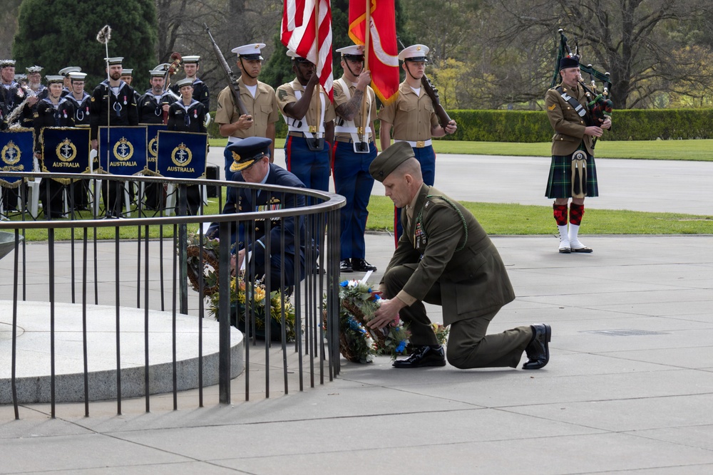 A formal military wreath-laying ceremony is taking place at what appears to be a memorial or tomb. Multiple military personnel from different nations are present, including Australian Navy band members, U.S. Marines in a color guard, and a Scottish bagpiper in traditional tartan kilt. Two officers are kneeling to place floral wreaths at a stone monument.