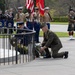 A formal military wreath-laying ceremony is taking place at what appears to be a memorial or tomb. Multiple military personnel from different nations are present, including Australian Navy band members, U.S. Marines in a color guard, and a Scottish bagpiper in traditional tartan kilt. Two officers are kneeling to place floral wreaths at a stone monument.