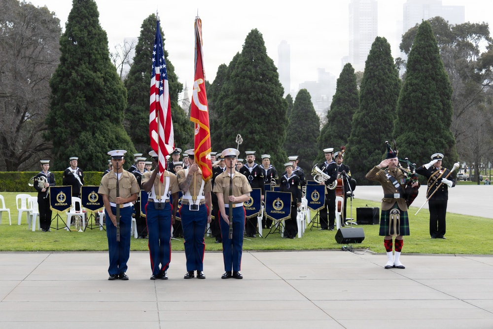 A joint military ceremony is taking place outdoors, featuring a U.S. Marine Corps color guard presenting the American flag and the Marine Corps flag. Behind them stands an Australian Navy band with members holding instruments and 'Australia'-labeled banners. A bagpiper in traditional Scottish Highland dress is visible to the right, saluting.