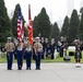 A joint military ceremony is taking place outdoors, featuring a U.S. Marine Corps color guard presenting the American flag and the Marine Corps flag. Behind them stands an Australian Navy band with members holding instruments and 'Australia'-labeled banners. A bagpiper in traditional Scottish Highland dress is visible to the right, saluting.