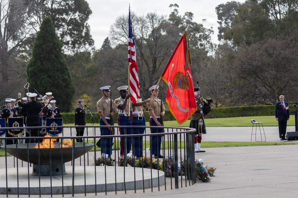 A formal military ceremony is taking place outdoors, featuring a U.S. Marine Corps color guard presenting the American flag and the Marine Corps flag alongside an eternal flame or ceremonial fire bowl. An Australian military band in dark uniforms is visible to the left, and a bagpiper in a kilt stands nearby, suggesting a joint U.S.-Australian commemorative event. A civilian in a suit observes from the right side of the frame.