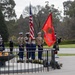 A formal military ceremony is taking place outdoors, featuring a U.S. Marine Corps color guard presenting the American flag and the Marine Corps flag alongside an eternal flame or ceremonial fire bowl. An Australian military band in dark uniforms is visible to the left, and a bagpiper in a kilt stands nearby, suggesting a joint U.S.-Australian commemorative event. A civilian in a suit observes from the right side of the frame.