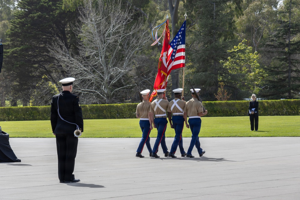 A U.S. Marine Corps color guard is performing a ceremonial parade on an outdoor plaza surrounded by manicured grounds and trees. Four Marines in dress uniform carry the American flag and Marine Corps colors while marching in formation. A senior officer in dress blues stands at attention observing the procession, with a civilian observer visible in the background.