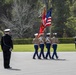 A U.S. Marine Corps color guard is performing a ceremonial parade on an outdoor plaza surrounded by manicured grounds and trees. Four Marines in dress uniform carry the American flag and Marine Corps colors while marching in formation. A senior officer in dress blues stands at attention observing the procession, with a civilian observer visible in the background.