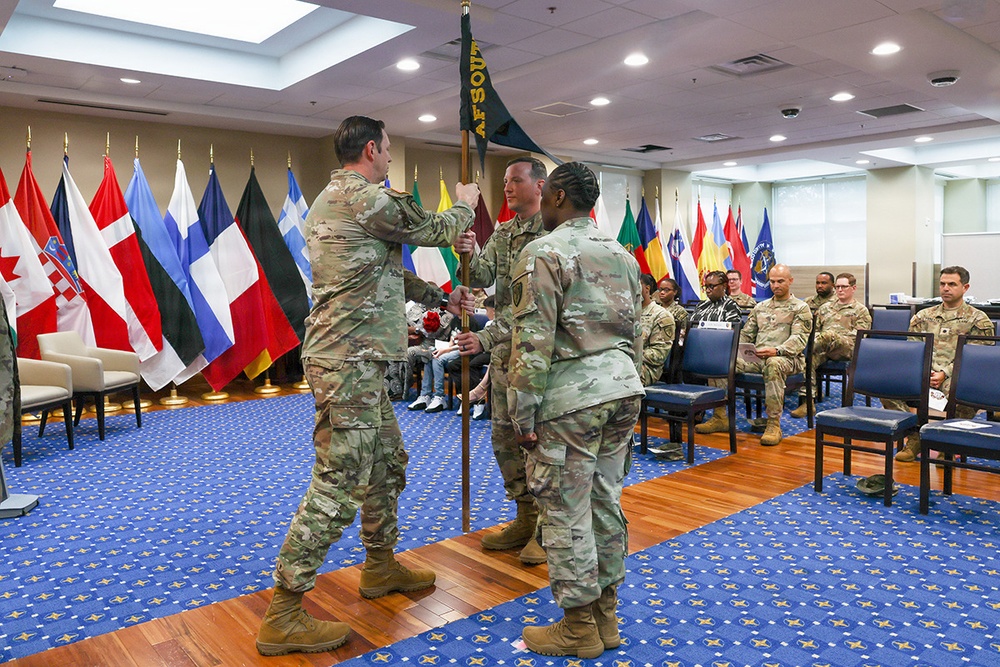 A military change of command or guidon ceremony is taking place indoors, with soldiers in U.S. Army OCP (Operational Camouflage Pattern) uniforms exchanging a unit guidon flag. The ceremony is conducted in a formal conference or ceremonial room adorned with multiple international and national flags along the walls. Seated military personnel serve as witnesses to the formal proceedings.