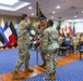 A military change of command or guidon ceremony is taking place indoors, with soldiers in U.S. Army OCP (Operational Camouflage Pattern) uniforms exchanging a unit guidon flag. The ceremony is conducted in a formal conference or ceremonial room adorned with multiple international and national flags along the walls. Seated military personnel serve as witnesses to the formal proceedings.