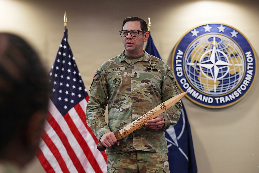 A U.S. Army soldier, identified by name tape as 'WELCH,' stands before a NATO Allied Command Transformation emblem and an American flag. He is holding a large wooden sword-shaped plaque engraved with the name '1SG Aaron M. Welch,' suggesting a military recognition or retirement ceremony. The setting appears to be an official NATO or U.S. military command facility.