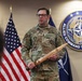 A U.S. Army soldier, identified by name tape as 'WELCH,' stands before a NATO Allied Command Transformation emblem and an American flag. He is holding a large wooden sword-shaped plaque engraved with the name '1SG Aaron M. Welch,' suggesting a military recognition or retirement ceremony. The setting appears to be an official NATO or U.S. military command facility.