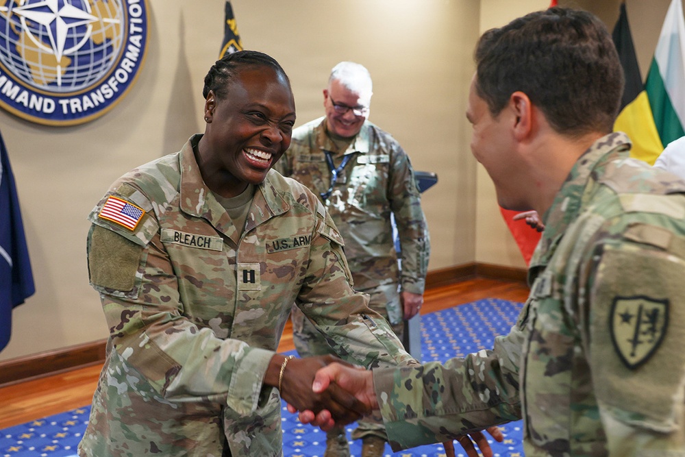 A U.S. Army soldier named BLEACH, wearing captain rank insignia, is shaking hands and smiling broadly with another soldier in a formal military setting. The scene appears to take place at NATO Allied Command Transformation headquarters, as indicated by the visible seal on the wall. A third soldier in the background observes the interaction with a smile.