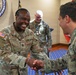 A U.S. Army soldier named BLEACH, wearing captain rank insignia, is shaking hands and smiling broadly with another soldier in a formal military setting. The scene appears to take place at NATO Allied Command Transformation headquarters, as indicated by the visible seal on the wall. A third soldier in the background observes the interaction with a smile.