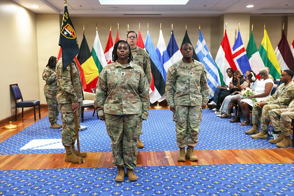 A formal U.S. Army military ceremony is taking place in an indoor venue decorated with multiple international NATO/allied nation flags. Several soldiers in OCP (Operational Camouflage Pattern) uniforms are standing at attention, with one soldier carrying a unit guidon/flag. Seated civilians and additional military personnel line the sides of the room, suggesting a promotion, change of command, or similar official ceremony.