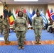 A formal U.S. Army military ceremony is taking place in an indoor venue decorated with multiple international NATO/allied nation flags. Several soldiers in OCP (Operational Camouflage Pattern) uniforms are standing at attention, with one soldier carrying a unit guidon/flag. Seated civilians and additional military personnel line the sides of the room, suggesting a promotion, change of command, or similar official ceremony.