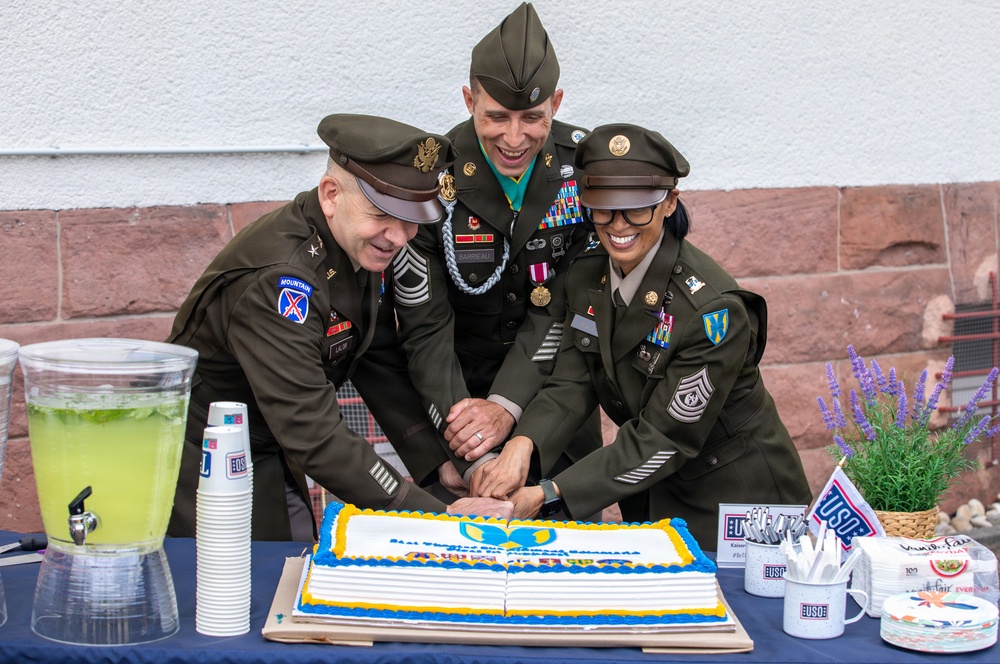 Three U.S. Army soldiers in dress green uniforms are jointly cutting a decorated cake at what appears to be a USO event or military ceremony. The cake features blue and gold frosting with a USO logo design, and the table is set with USO-branded merchandise and refreshments. All three individuals are smiling and appear to be in a celebratory mood.