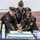 Three U.S. Army soldiers in dress green uniforms are jointly cutting a decorated cake at what appears to be a USO event or military ceremony. The cake features blue and gold frosting with a USO logo design, and the table is set with USO-branded merchandise and refreshments. All three individuals are smiling and appear to be in a celebratory mood.