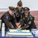 Three U.S. Army soldiers in dress green uniforms are jointly cutting a decorated cake at what appears to be a USO event or military ceremony. The cake features blue and gold frosting with a USO logo design, and the table is set with USO-branded merchandise and refreshments. All three individuals are smiling and appear to be in a celebratory mood.