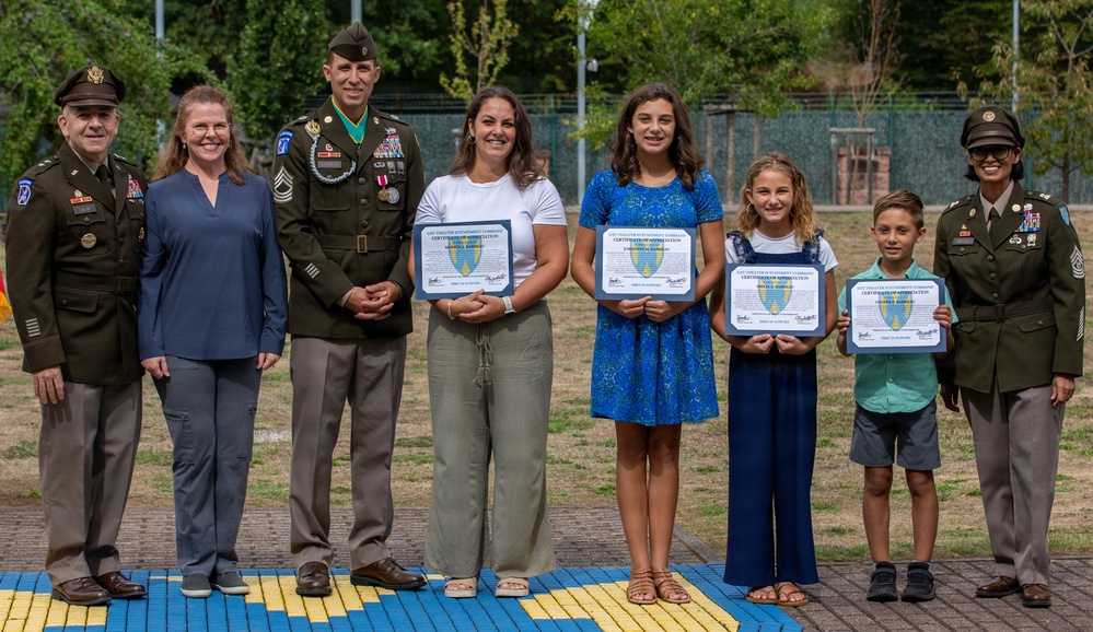 A military awards ceremony featuring a group of seven individuals posing outdoors. Three military personnel in U.S. Army uniforms flank a civilian family of four, who are holding framed Certificates of Appreciation from what appears to be the U.S. Army Theater Sustainment Command. The ceremony appears to be recognizing the family members, including children, for their contributions or support.