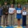 A military awards ceremony featuring a group of seven individuals posing outdoors. Three military personnel in U.S. Army uniforms flank a civilian family of four, who are holding framed Certificates of Appreciation from what appears to be the U.S. Army Theater Sustainment Command. The ceremony appears to be recognizing the family members, including children, for their contributions or support.