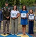 A military awards ceremony featuring a group of seven individuals posing outdoors. Three military personnel in U.S. Army uniforms flank a civilian family of four, who are holding framed Certificates of Appreciation from what appears to be the U.S. Army Theater Sustainment Command. The ceremony appears to be recognizing the family members, including children, for their contributions or support.