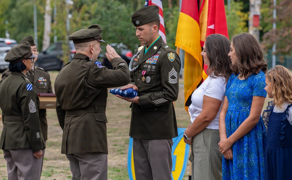 A military retirement or awards ceremony is taking place outdoors, where a senior officer is saluting and presenting a folded American flag to a decorated non-commissioned officer (NCO). The NCO, wearing dress greens with numerous medals and a green beret cord, stands alongside what appears to be his family — a woman and two children. Military color guard members and unit flags are visible in the background.