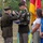 A military retirement or awards ceremony is taking place outdoors, where a senior officer is saluting and presenting a folded American flag to a decorated non-commissioned officer (NCO). The NCO, wearing dress greens with numerous medals and a green beret cord, stands alongside what appears to be his family — a woman and two children. Military color guard members and unit flags are visible in the background.