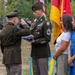 A military retirement or awards ceremony is taking place outdoors, where a senior officer is saluting and presenting a folded American flag to a decorated non-commissioned officer (NCO). The NCO, wearing dress greens with numerous medals and a green beret cord, stands alongside what appears to be his family — a woman and two children. Military color guard members and unit flags are visible in the background.
