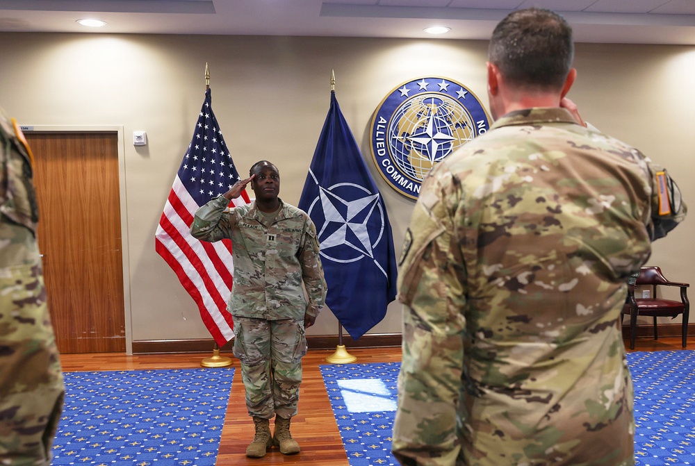 A U.S. Army soldier in OCP (Operational Camouflage Pattern) uniform is rendering a salute during what appears to be a military ceremony at a NATO Allied Command facility. The setting includes both the American flag and the NATO flag, with the NATO Allied Command emblem prominently displayed on the wall. At least two other uniformed soldiers are present, one facing the saluting soldier.