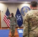 A U.S. Army soldier in OCP (Operational Camouflage Pattern) uniform is rendering a salute during what appears to be a military ceremony at a NATO Allied Command facility. The setting includes both the American flag and the NATO flag, with the NATO Allied Command emblem prominently displayed on the wall. At least two other uniformed soldiers are present, one facing the saluting soldier.