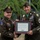 Two U.S. Army soldiers pose together holding a framed Certificate of Appreciation during what appears to be a military retirement or awards ceremony. The officer on the left, wearing two-star Major General rank insignia and a 10th Mountain Division patch, is presenting the certificate to a Master Sergeant on the right who is wearing the Medal of Honor around his neck. The certificate is addressed to Master Sergeant Caleb Barrieau, United States Army.